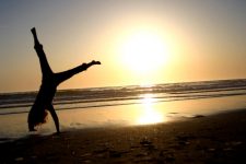 Foto de uma moça dando "estrelinha" na praia no pôr do sol.
