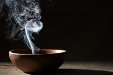 A foto mostra um cerâmica marrom, com uma fumaça de incenso saindo dele. A foto está com o fundo escuro e dá bastante destaque para a fumaça. O incenso revigora seu espírito