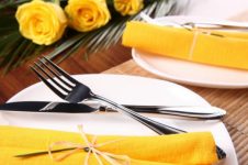 A foto mostra pratos brancos em uma mesa de madeira, com destaque para um dos pratos com os talheres cruzados e um guardanapo de pano amarelo. Há uma rosa amarela ao fundo. A foto ilustra a importancia da mastigação no emagrecimento