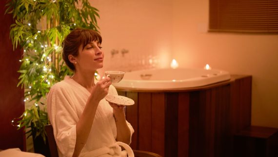 Mulher relaxando em um ambiente de spa, vestindo roupão branco e saboreando uma xícara de chá, com banheira ao fundo, luzes suaves e velas acesas, transmitindo paz e autocuidado.